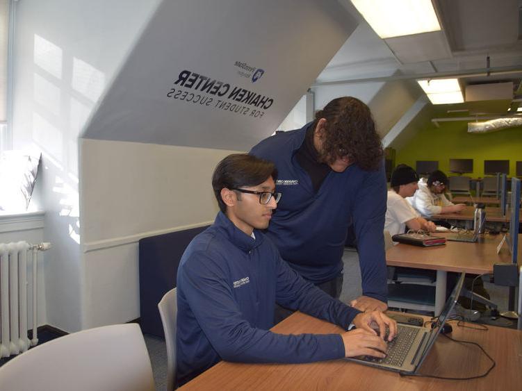 Person standing and person sitting and looking at a laptop